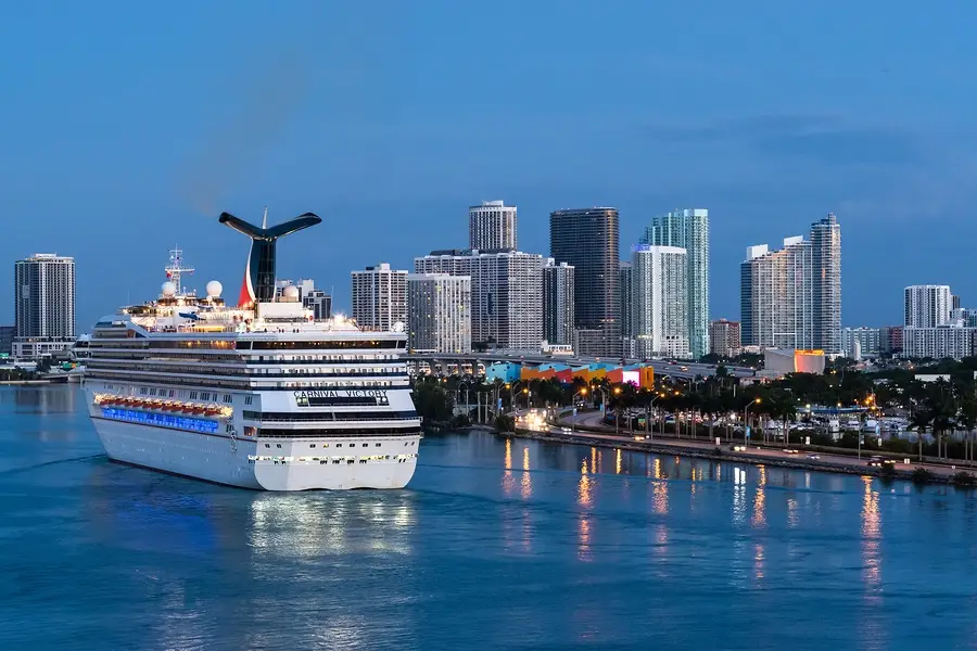 Cruise ship Carnival Victory navigating near Port Miami, with the Miami skyline featuring modern skyscrapers in the background, highlighting upcoming renovations and impact on the city's cruise industry.