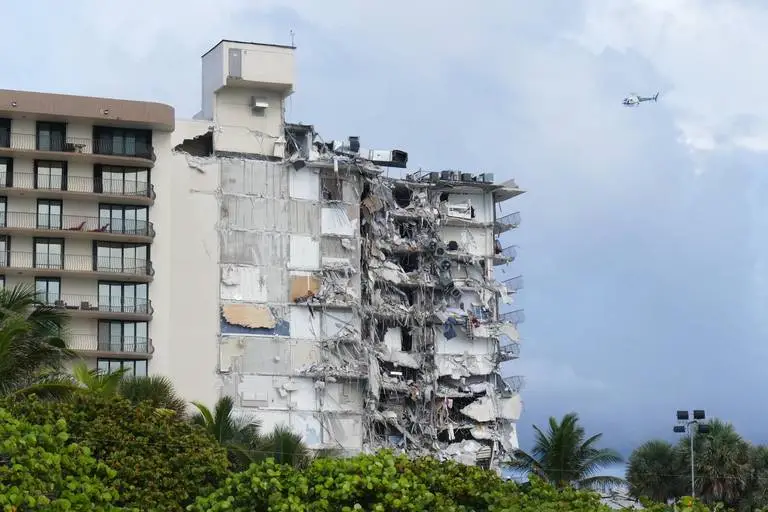 Damaged Champlain Towers South condominium in Surfside, showing significant structural collapse and debris, with a helicopter overhead, related to recent tragic events and ongoing legal actions.