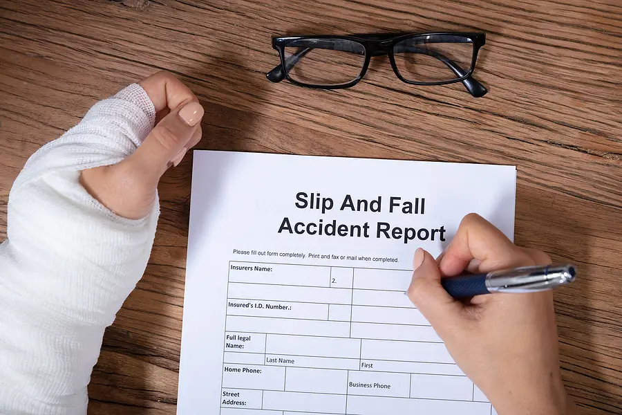 Person with a bandaged hand filling out a slip and fall accident report on a wooden table, with glasses in the background, emphasizing personal injury documentation.