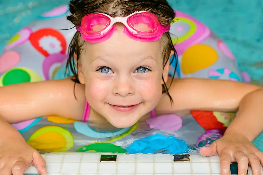 Young girl with pink swim goggles smiling in a colorful inflatable ring, enjoying swimming in a pool, reflecting themes of water safety and drowning prevention in Florida.
