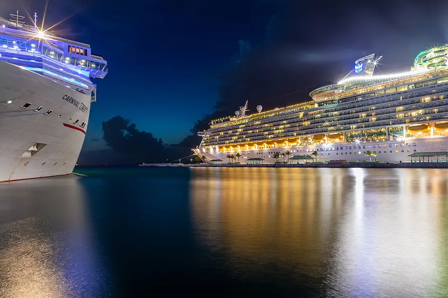 Carnival cruise ship and Royal Caribbean cruise ship docked at night, illuminated reflections in the water, showcasing the cruise industry.