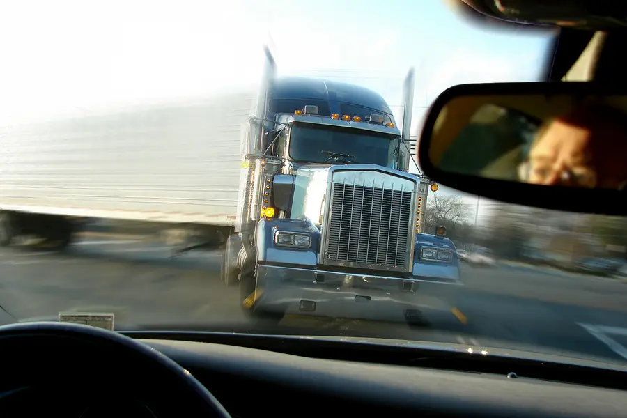 Close-up view of a large truck approaching head-on on a road, with a blurred background and a glimpse of a driver's face in the rearview mirror, highlighting the dangers of truck accidents on Florida roads.