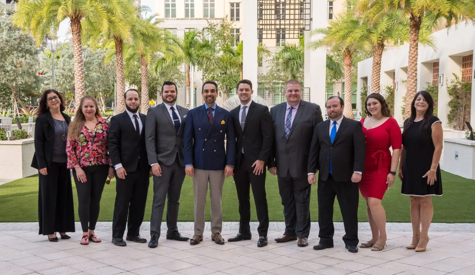 Group of professional individuals in formal attire posing outdoors, representing Aronfeld Trial Lawyers, with palm trees and modern architecture in the background.