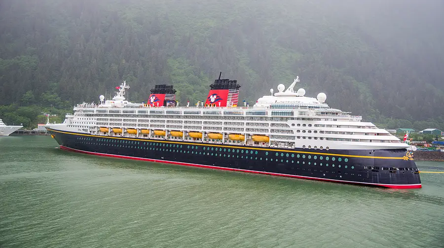Disney Cruise Line ship in port, showcasing iconic red funnels and colorful exterior, amidst lush green hills, relevant to upcoming cruise announcements.