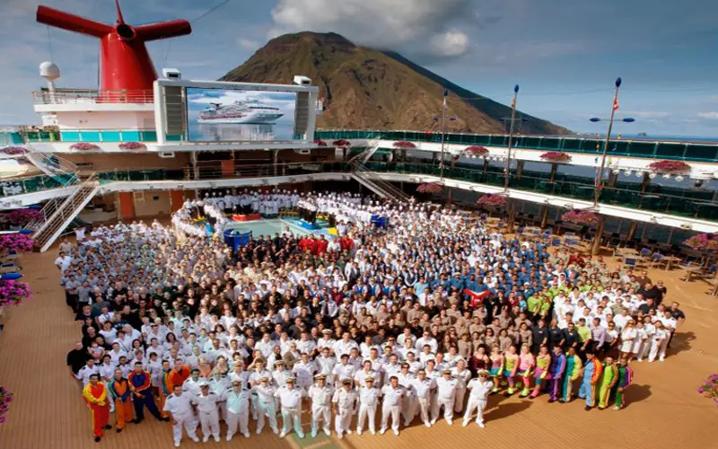 Carnival cruise ship crew members posing on deck with mountain backdrop, showcasing diverse uniforms and team spirit, relevant to maritime law and cruise safety discussions.