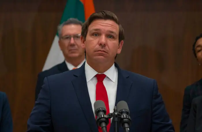 Florida Governor Ron DeSantis speaking at a podium with microphones, addressing legal actions against the CDC regarding cruise operations, with flags in the background.