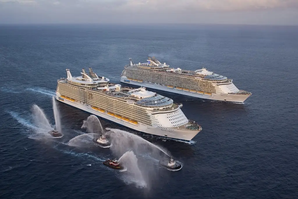 Two of the world's largest cruise ships at sea, surrounded by tugboats spraying water, showcasing the scale and luxury of modern maritime travel.