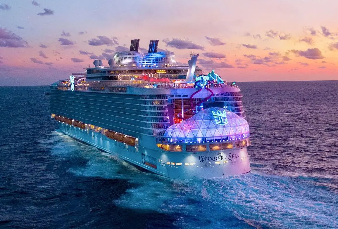 Aerial view of the Wonder of the Seas cruise ship at sunset, showcasing vibrant lighting and ocean waves, emphasizing its status as the world's largest cruise ship.