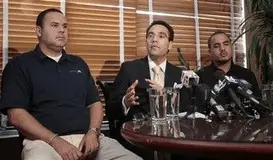 Three men seated at a table during a press conference, discussing workplace injuries related to traffic accidents, with microphones and water glasses present.
