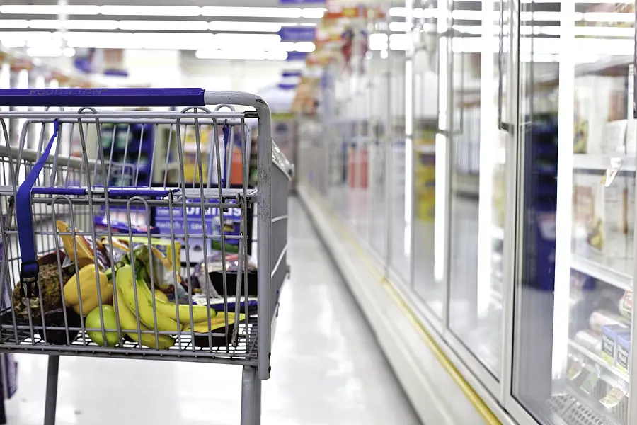 Grocery shopping cart filled with fresh produce, including bananas and apples, in a well-lit supermarket aisle, illustrating the importance of safety while shopping during the busy Thanksgiving season.