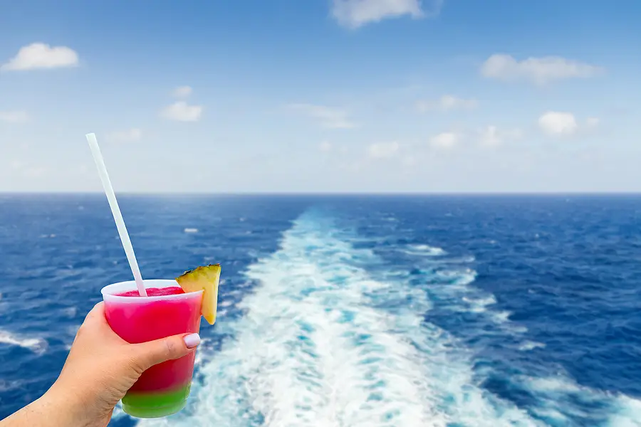 Hand holding a colorful cocktail with a straw, overlooking the ocean and wake of a cruise ship under a blue sky, symbolizing leisure and relaxation on a cruise vacation.