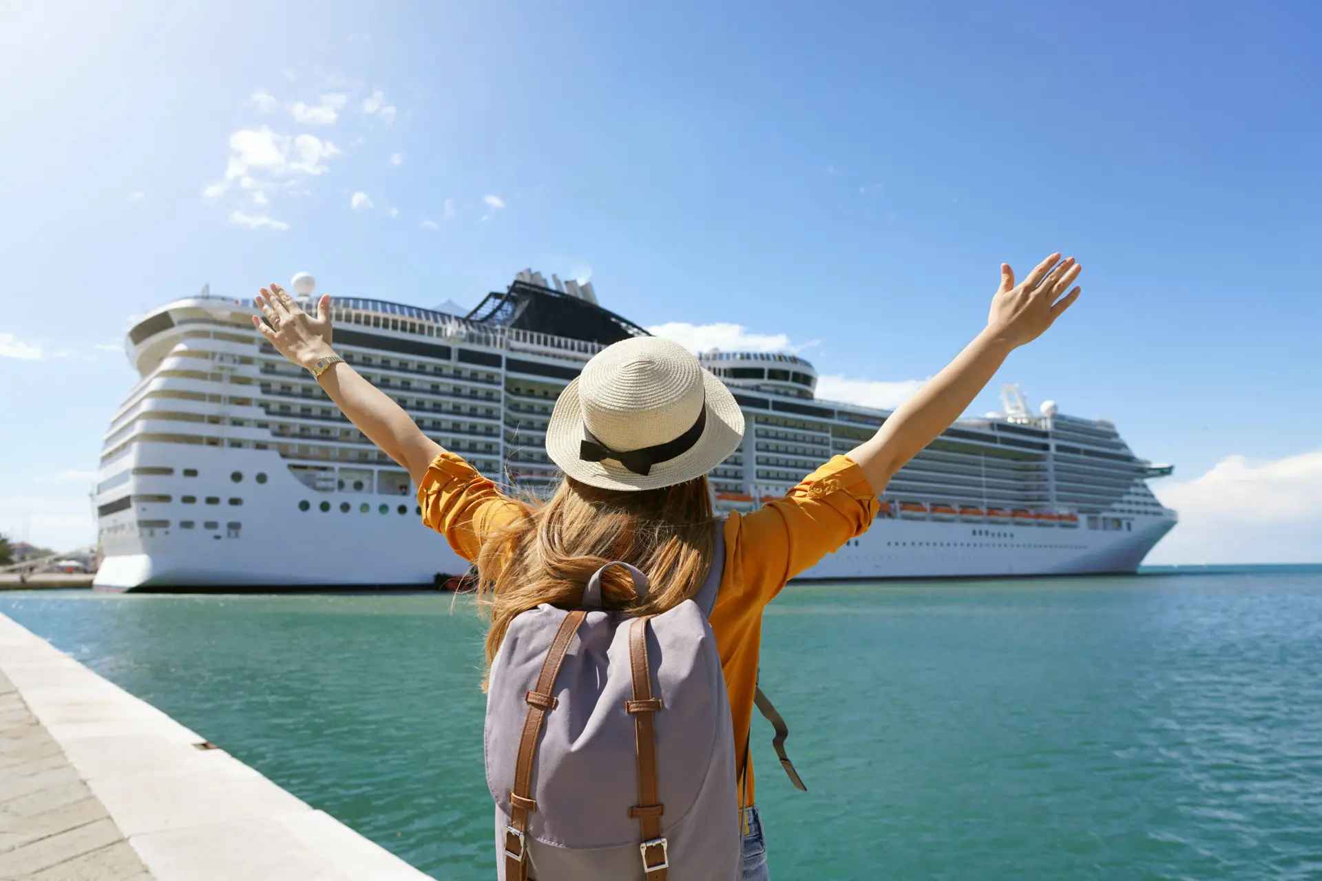 Woman in a yellow shirt and straw hat raising arms in excitement near a large cruise ship in the Port of Zeebrugge, Belgium, emphasizing cruise travel and maritime experiences.