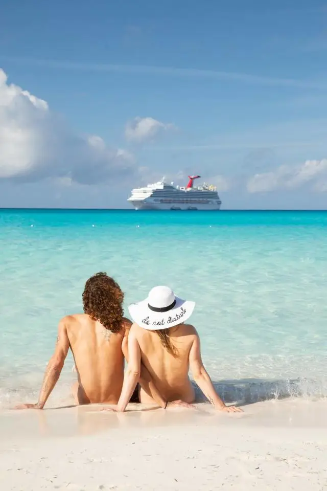 Couple sitting on the beach in the foreground, enjoying a nude cruise experience, with a Carnival cruise ship in the background against a clear blue sky and turquoise water.