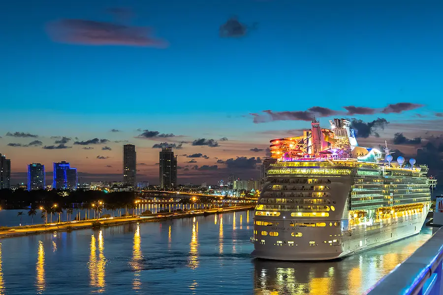 Miami skyline at dusk with Royal Caribbean cruise ship, Hero of the Seas, illuminated and docked, reflecting lights on water, showcasing vibrant city life and maritime adventure.